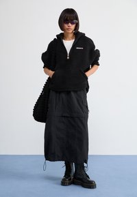 Haut à capuche en polaire noire avec une demi-fermeture éclair et une poche avant, associé à une jupe cargo noire et des bottes épaisses à lacets noires. Sac noir texturé.