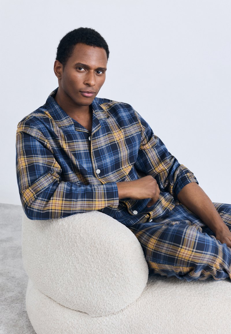 Blå och gul rutig pyjamas i mjukt tyg, med en knappjacket och matchande byxor, bärs medan personen sitter på en texturerad krämfärgad stol.