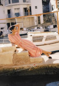 Femme en robe à motifs se détendant sur des marches en pierre près d'un quai, avec de petits bateaux et des bâtiments en arrière-plan.