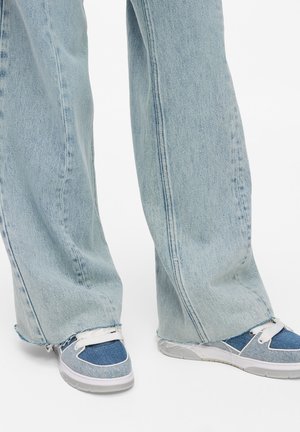Svijetloplave jeans hlače širokog kroja s rasparanim rubovima, uparene s višebojnim plavim tenisicama koje imaju bijeli potplat i vezice. Glatka jeans tekstura.