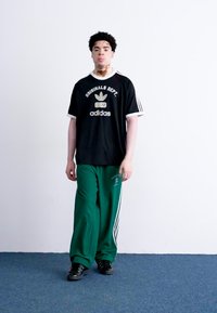 Črna adidas majica s belimi detajli in logotipom, v kombinaciji z zelenimi trenirkami, ki imajo bele črte, oblečeno s črnimi supergami na modrem preprogu.