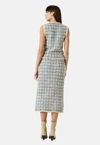 Vrouw in een mouwloze blauw en beige gestructureerde midi-jurk met franjes, gecombineerd met beige hakken tot de enkel, staand met haar rug naar de camera.