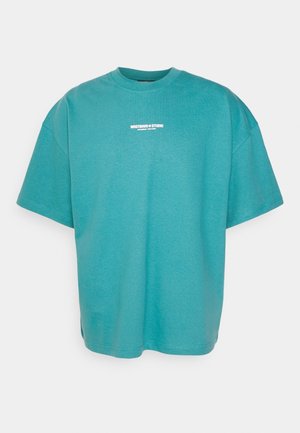 T-shirt oversize bleu sarcelle à manches courtes, col rond, avec un petit texte blanc centré sur la poitrine.