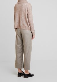 Vaaleanruskea, ylisuuri neulepaita, jossa on korkea kaulus; vaaleanharmaat, leveät housut; mustat, litteät kengät, joissa on koristeellinen yksityiskohta.