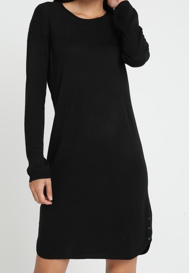 Robe en maille noire à manches longues avec encolure ronde, détails de boutons près de l'ourlet, portée par une personne debout devant un fond uni.
