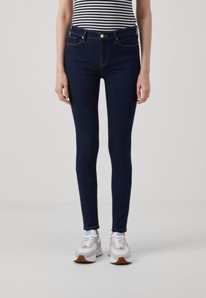 Kvinde iført mørkeblå skinny jeans, hvide og blå sneakers samt en sort-hvid stribet ærmeløs top, stående foran en ensfarvet baggrund.