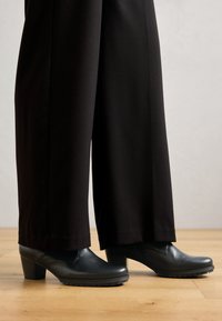 Des chaussures à talons noirs avec un bout arrondi et une semelle en caoutchouc texturé, associées à un pantalon large noir, le tout sur un sol en bois clair.