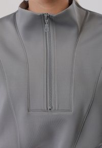 Gros plan sur un vêtement gris à col montant avec une fermeture éclair verticale centrale et des lignes de couture visibles sur le tissu.