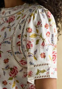 Biała koszula w kwiaty z bufiastymi rękawami, zdobiona kolorowymi kwiatami oraz misternymi wstawkami koronkowymi wzdłuż dekoltu i rękawów.