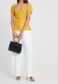 Blusa gialla a pois con maniche a palloncino e bottoni, abbinata a pantaloni bianchi a gamba larga. Tiene in mano una borsa nera con manico di legno.