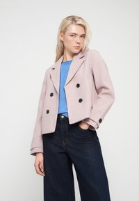 Ρόζ διπλόπατο σακάκι από μάλλινο μείγμα, με μαύρα κουμπιά, δομημένες πέτσες και φαρδιά μανίκια. Φορεμένο πάνω από μπλε τοπ και σκούρο τζιν.