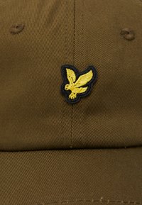 Barna pamut sapka kiemelkedő sárga sas logóval, fekete kontúrral hímezve. Texturált anyag és hat paneles kialakítás.