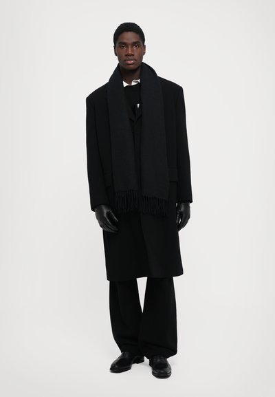 Lång svart kappa med hög krage, ihop med en svart halsduk och handskar. Svarta byxor och skor kompletterar outfiten. Enkel, monokromatisk design.