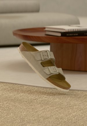 Beige suede sandal with two adjustable buckles, cork footbed, and white sole floating above a textured beige rug near a wooden coffee table.