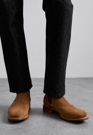 Pruunid sueded Chelsea saapad elastsete külgpaneelide ja virnastatud puidust pealsetega, kantud tumedate, veidi tekstureeritud pükstega.