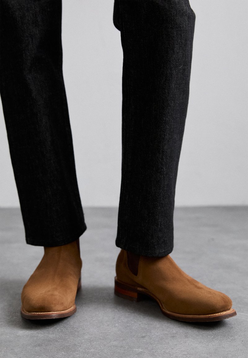 Barna velúr Chelsea csizma, elasztikus oldalsó panelekkel és rakásolt fa sarokkal, sötét, enyhén texturált nadrággal viselve.