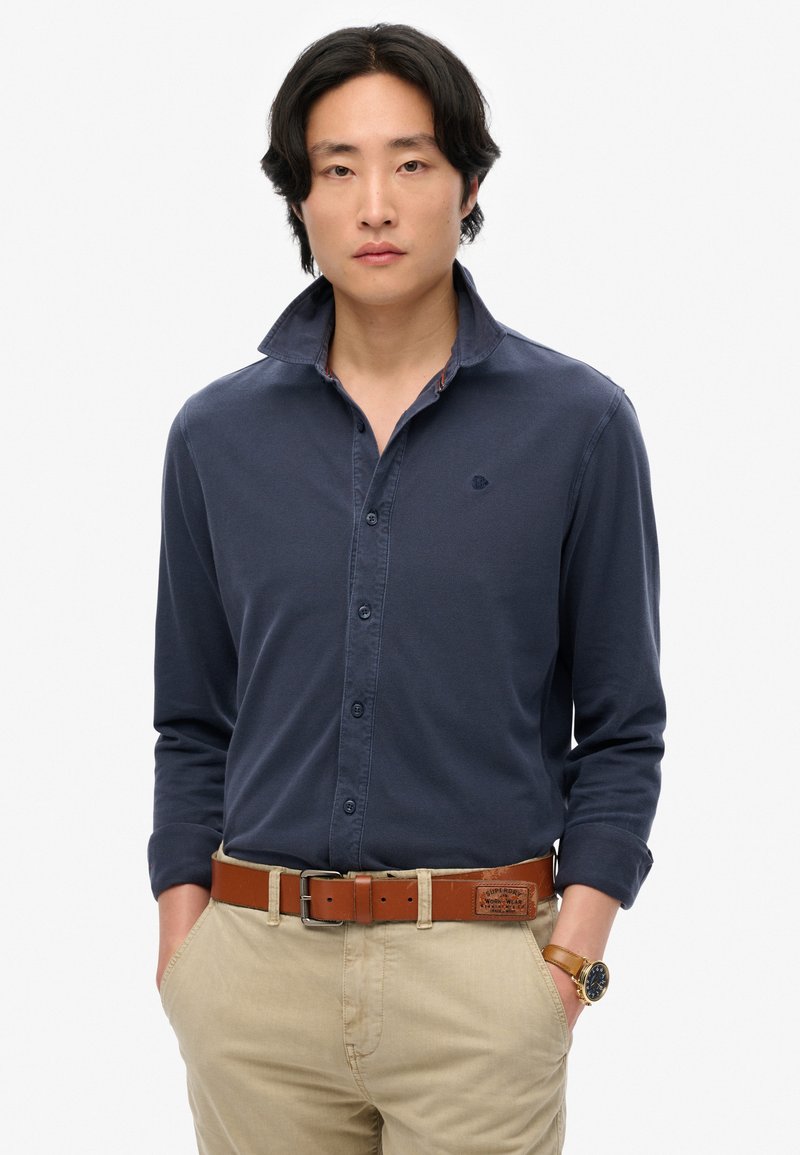 Marineblauw polo-shirt met lange mouwen en een button-down kraag, met subtiele logo-borduring en een bruine leren riem met een rechthoekige gesp.