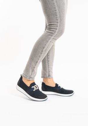 Marineblaue Slipper aus strukturiertem Stoff, mit einer dekorativen Schnalle und weißer Sohle. Tragen mit grauen, enganliegenden Jeans.