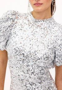 Zilveren paillettenjurk met pofmouwen, ronde halslijn en getailleerde pasvorm. Sprankelende textuur die licht weerkaatst met verschillende paillettenformaten.