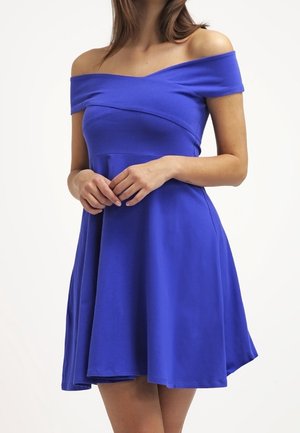 Femme portant une robe bleue épaules dénudées avec un corsage ajusté et une jupe évasée, les mains doucement croisées devant elle.