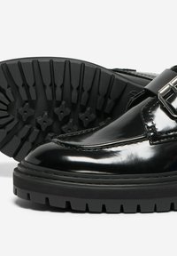Chaussures en cuir verni noir avec une finition brillante, semelle en caoutchouc noir épaisse et un accent de boucle décoratif sur la partie supérieure.