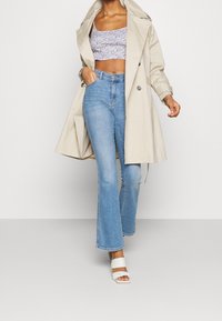 Beige trenchcoat över en lavendelfärgad crop top, tillsammans med ljusblå utsvängda jeans och vita blockklackade sandaler.