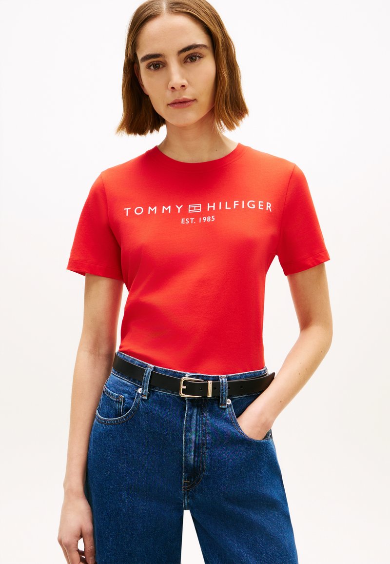 Maglietta rossa in cotone con maniche corte e logo bianco "TOMMY HILFIGER EST. 1985", abbinata a jeans di denim blu e cintura nera.