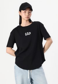 Svart kortermet t-skjorte med rund hem, med en hvit brodert "GAP"-logo på brystet. Brukt sammen med lyseblå jeans.