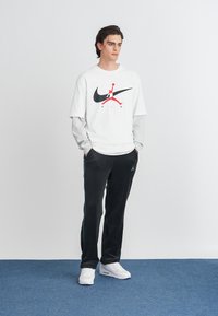 Balta medvilninė marškinėliai su juodu ir raudonu Nike Air logotipu, apsivilkti pilku ilgu rankovėmis marškinėliu. Juodos sportinės kelnės su mėlynu šonu, baltos sportinės bateliai.