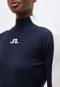 Κοντινή λήψη ενός ατόμου που φοράει μπλε ναυτικό και άσπρο πουλόβερ με ψηλό γιακά και μικρό λευκό λογότυπο στο στήθος.