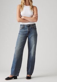 Femme debout, bras croisés, portant un haut blanc sans manches, un jean bleu taille haute et des ballerines noires sur fond gris.