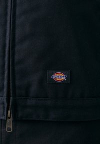 Black fabric workwear with a zippered closure. Features a rectangular Dickies logo patch in color on the right side. Textured canvas material.