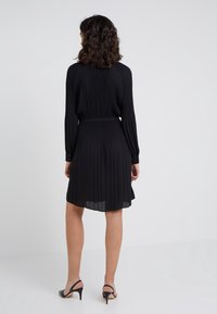 Robe noire plissée à manches longues, taille cintrée et jupe mi-longue. Texture lisse, tissu doux, associée à des escarpins à talon ouverts noirs.