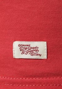 Rotes Stoffetikett mit weißer Naht. Das Label zeigt "Genuine Dry Goods & Supply" in roter Schreibschrift auf einem cremefarbenen Hintergrund. Originale Textur sichtbar.