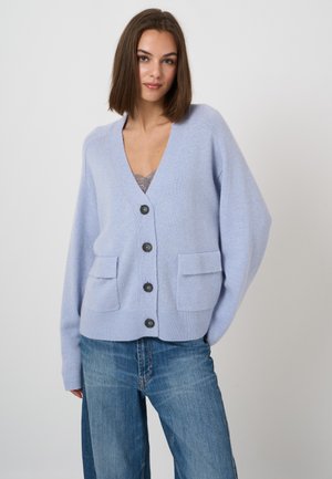 Young woman wearing a light blue buttoned cardigan with front pockets and blue jeans, standing against a plain white background.