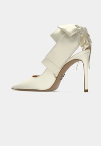 Escarpin blanc en satin à talon aiguille avec un bout pointu, doté d'une large sangle et d'un grand nœud à l'arrière, avec une semelle classique couleur marron clair.