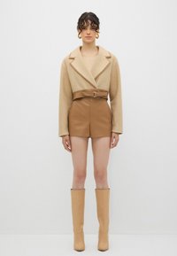 Beige uldblanding oversized jakke med bælte i taljen, som er parret med brune faux læder højtaljede shorts og knæhøje beige støvler.