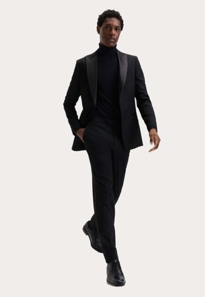 Mężczyzna idący w czarnym golfie, dopasowanej marynarce, spodniach i skórzanych eleganckich butach na jasnym, jednolitym tle.