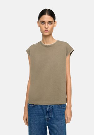 Młoda kobieta z ciemnymi, spiętymi włosami, ubrana w bezrękawnik oliwkowej zieleni i niebieskie dżinsy, stojąca na tle jednolitego jasnego tła.