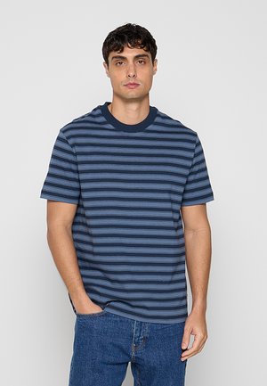 Mężczyzna o ciemnych kręconych włosach, ubrany w niebiesko-granatowy poziomo paskowany T-shirt i niebieskie dżinsy, stojący na tle jednolitego szarego tła.