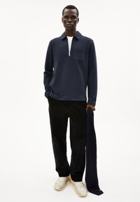 Navy zippered polo shirt with a chest pocket, black corduroy pants, and multi-colored sneakers. Holding a dark navy scarf.