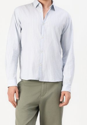 Mężczyzna ubrany w jasnoniebieską koszulę w paski z długim rękawem na guziki i oliwkowe spodnie, stojący na białym tle.