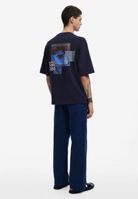 Camiseta de gran tamaño en azul marino con patrones geométricos en azul y marrón en la parte posterior, hecha de una tela suave. Combinada con pantalones anchos azules y sandalias negras.