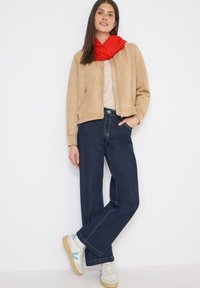 Femme debout contre un mur blanc, portant une veste beige, une écharpe rouge, un haut clair, un jean large sombre et des baskets blanches avec des accents bleus.