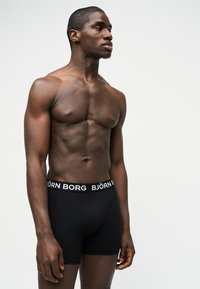 Καλοντυμένος άνδρας με σκούρο δέρμα που φοράει μαύρα boxer Björn Borg, όρθιος, στραμμένος ελαφρώς προς τα αριστερά, με φόντο απλό και ανοιχτόχρωμο.