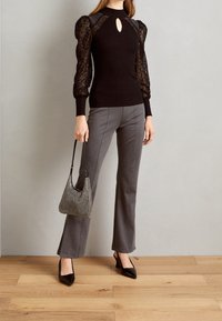 Pull noir à côtes avec des manches bouffantes en dentelle et un décolleté en goutte d'eau, associé à un pantalon gris évasé et des escarpins noirs à bout pointu, tenant un sac à main métallique.