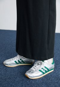 Chaussures de sport argentées avec des rayures vertes, des lacets blancs et une semelle en gomme. Portées avec un vêtement noir à côtes.