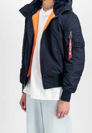 Marineblå bomberjakke med orange indre, pelsforet hætte, sølvlynlås og en rød mærkat med teksten "FJERN FØR FLYVNING" på ærmet.