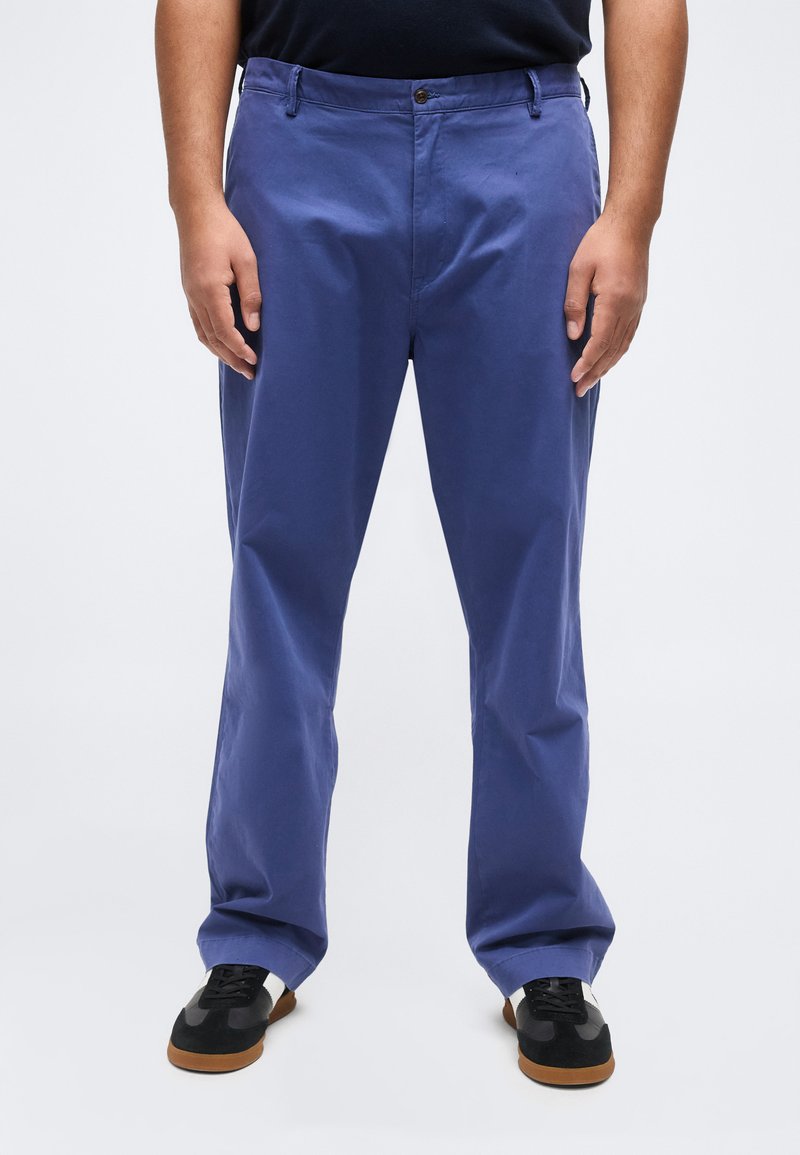 Άνδρας που φορά μπλε casual παντελόνια και μαύρα-λευκά αθλητικά παπούτσια, στέκεται με τα χέρια χαλαρά στο πλάι, μπροστά σε απλό φόντο.