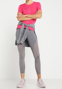 Rosa kortärmad skjorta med texten "JUST", grå leggings med nätpaneler och ljusgrå snören. Modellen poserar med en grå sweatshirt knuten vid midjan.